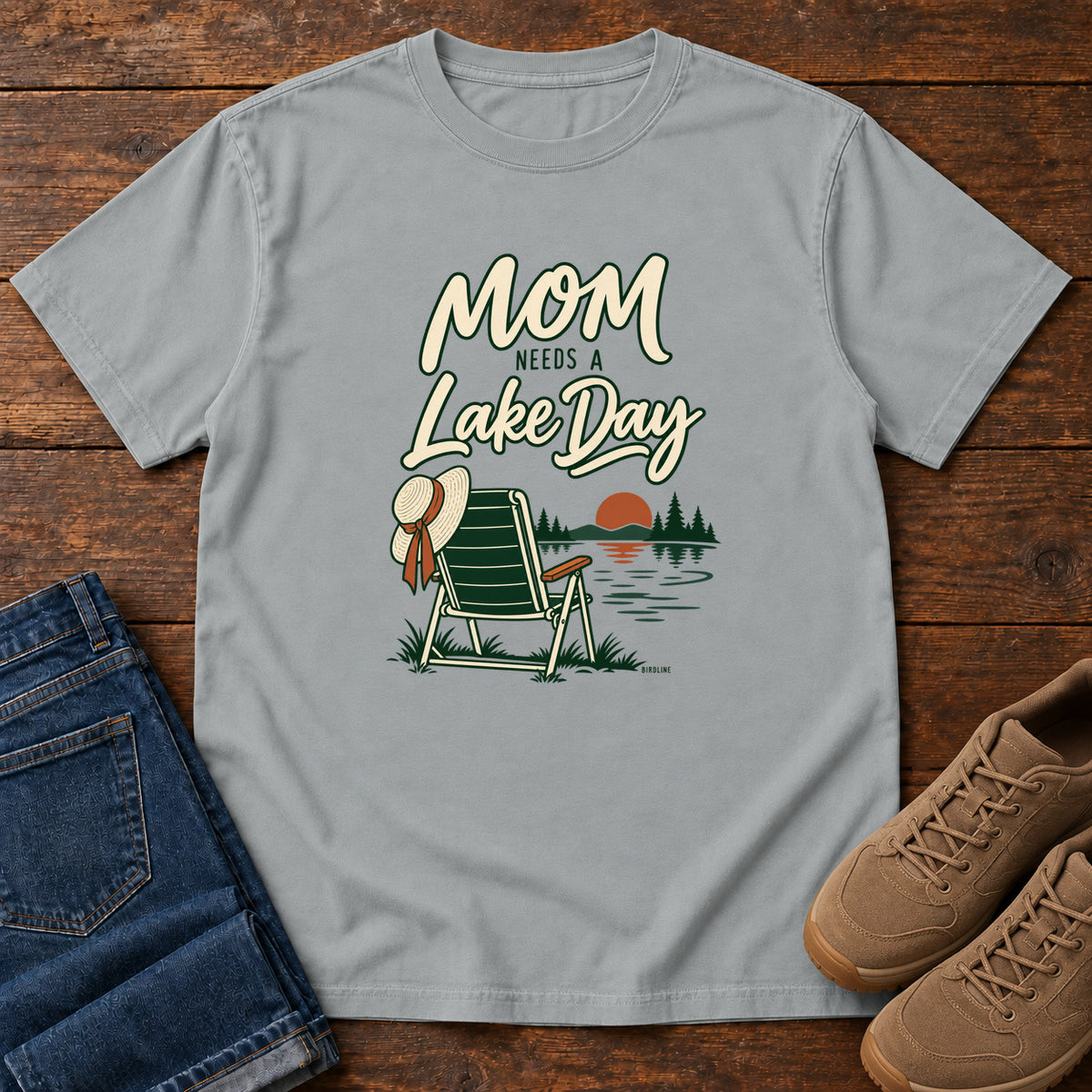 Granite Mom Needs a Lake Day T-Shirt with a striped folding chair, sunhat and lakeside sunset illustration, front flat lay on wood.