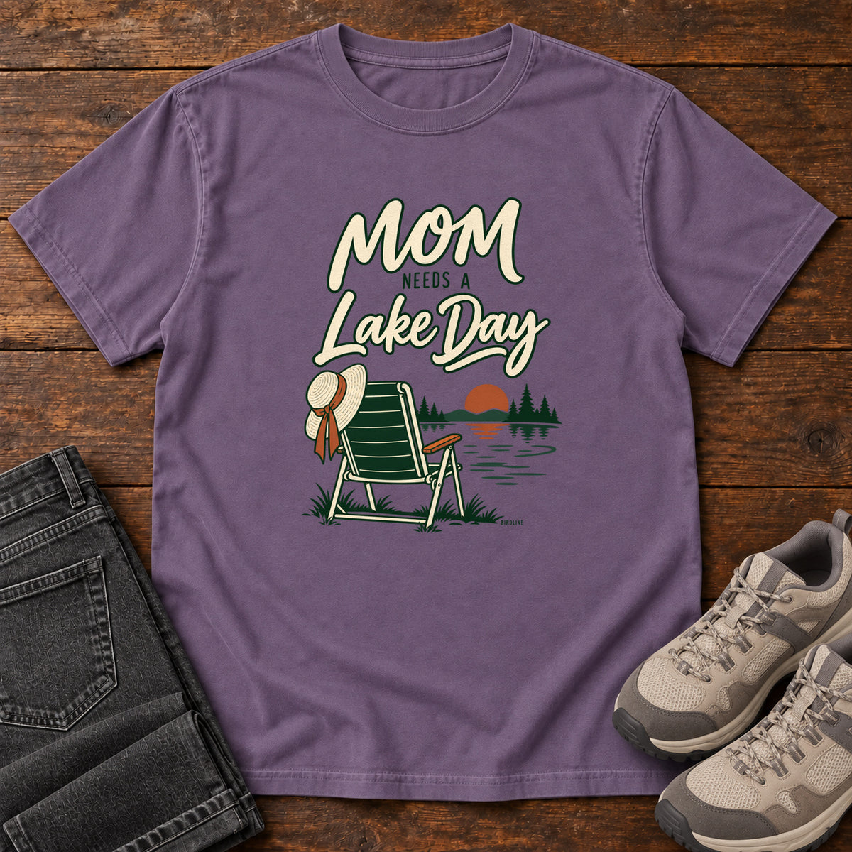 Grape Mom Needs a Lake Day T-Shirt with a striped folding chair, sunhat and lakeside sunset illustration, front flat lay on wood.