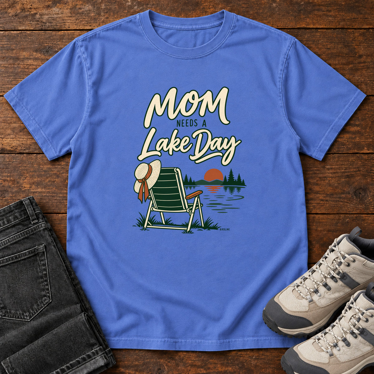 Mystic Blue Mom Needs a Lake Day T-Shirt with a striped folding chair, sunhat and lakeside sunset illustration, front flat lay on wood.