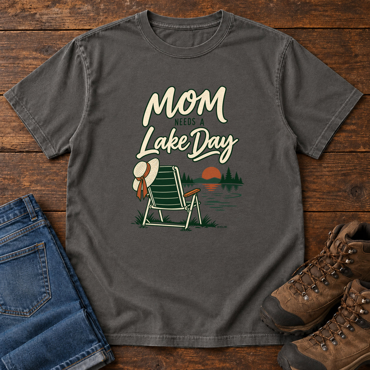 Pepper Mom Needs a Lake Day T-Shirt with a striped folding chair, sunhat and lakeside sunset illustration, front flat lay on wood.