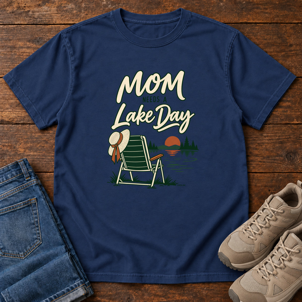 True Navy Mom Needs a Lake Day T-Shirt with a striped folding chair, sunhat and lakeside sunset illustration, front flat lay on wood.