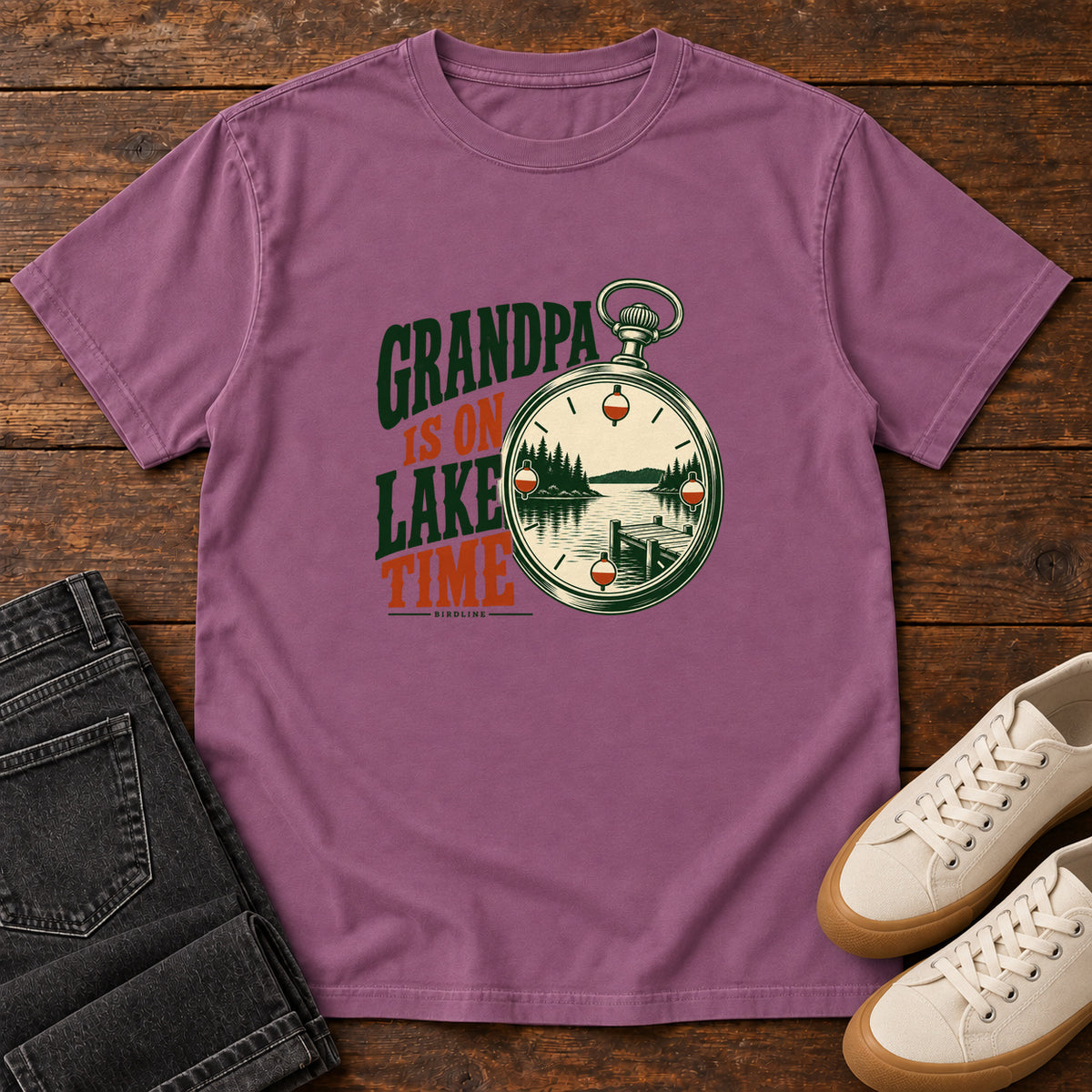 Berry Grandpa Is on Lake Time T-Shirt with a pocket watch framing a lake and dock beside green and orange lettering; front flat lay on wood.