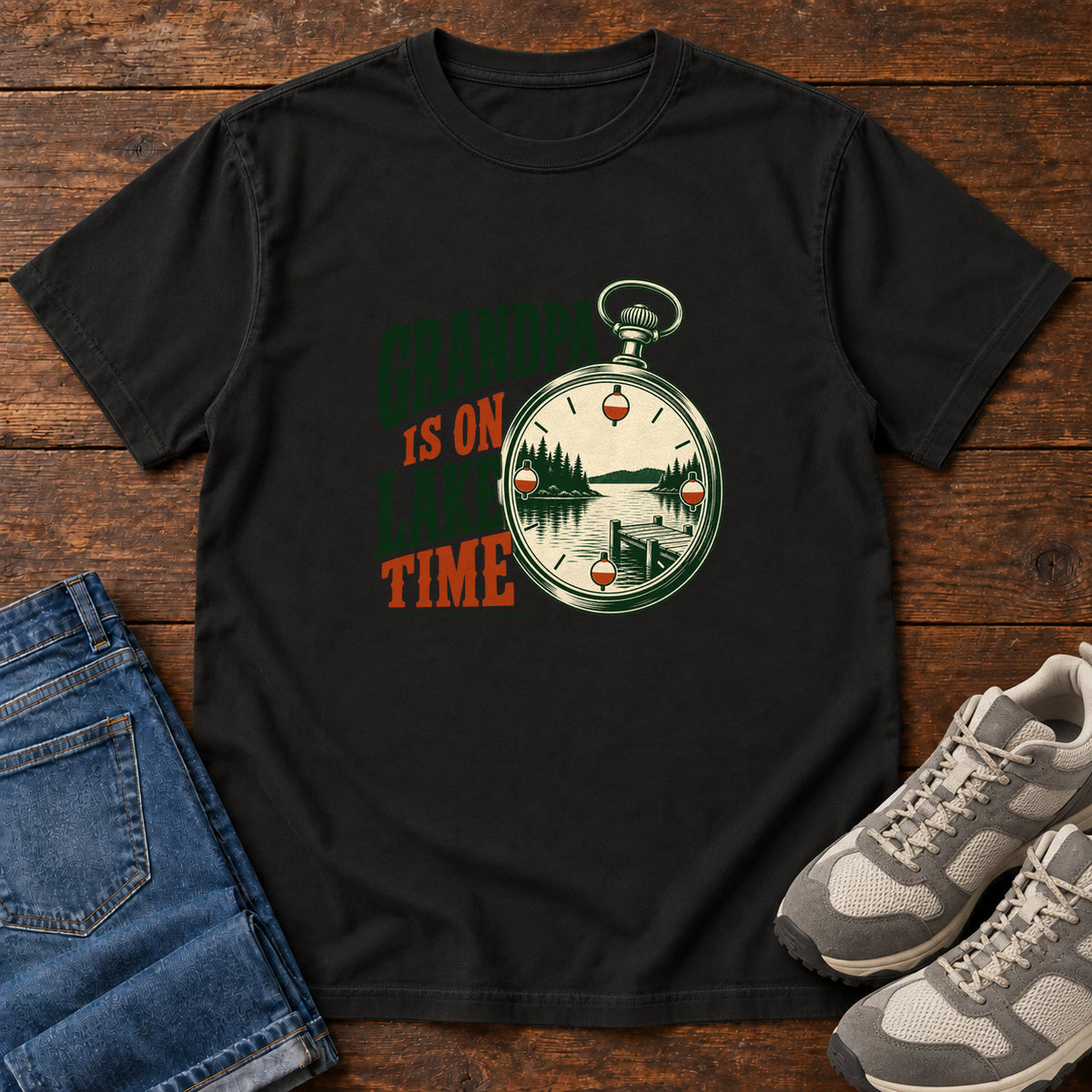 Black Grandpa Is on Lake Time T-Shirt with a pocket watch framing a lake and dock beside green and orange lettering; front flat lay on wood.