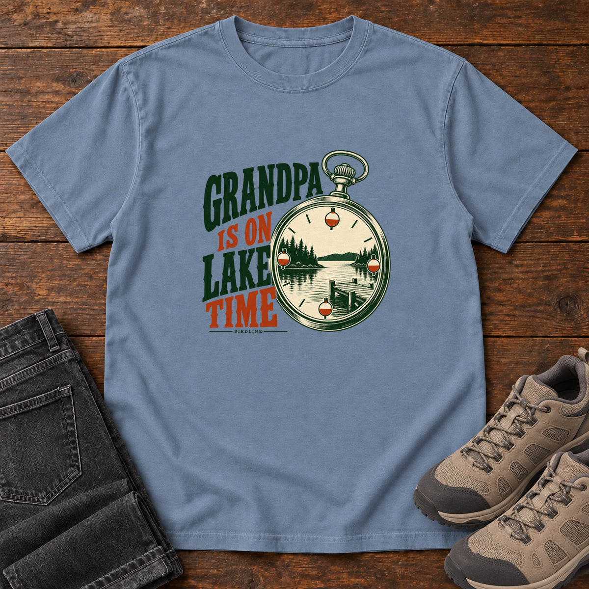 Blue Jean Grandpa Is on Lake Time T-Shirt with a pocket watch framing a lake and dock beside green and orange lettering; front flat lay on wood.