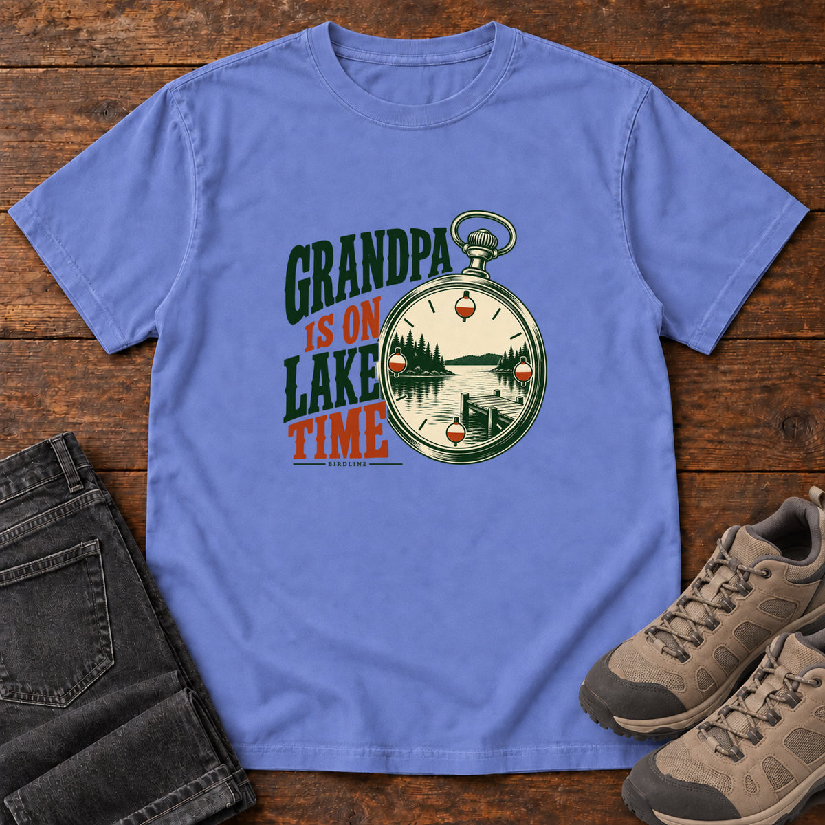 Flo Blue Grandpa Is on Lake Time T-Shirt with a pocket watch framing a lake and dock beside green and orange lettering; front flat lay on wood.