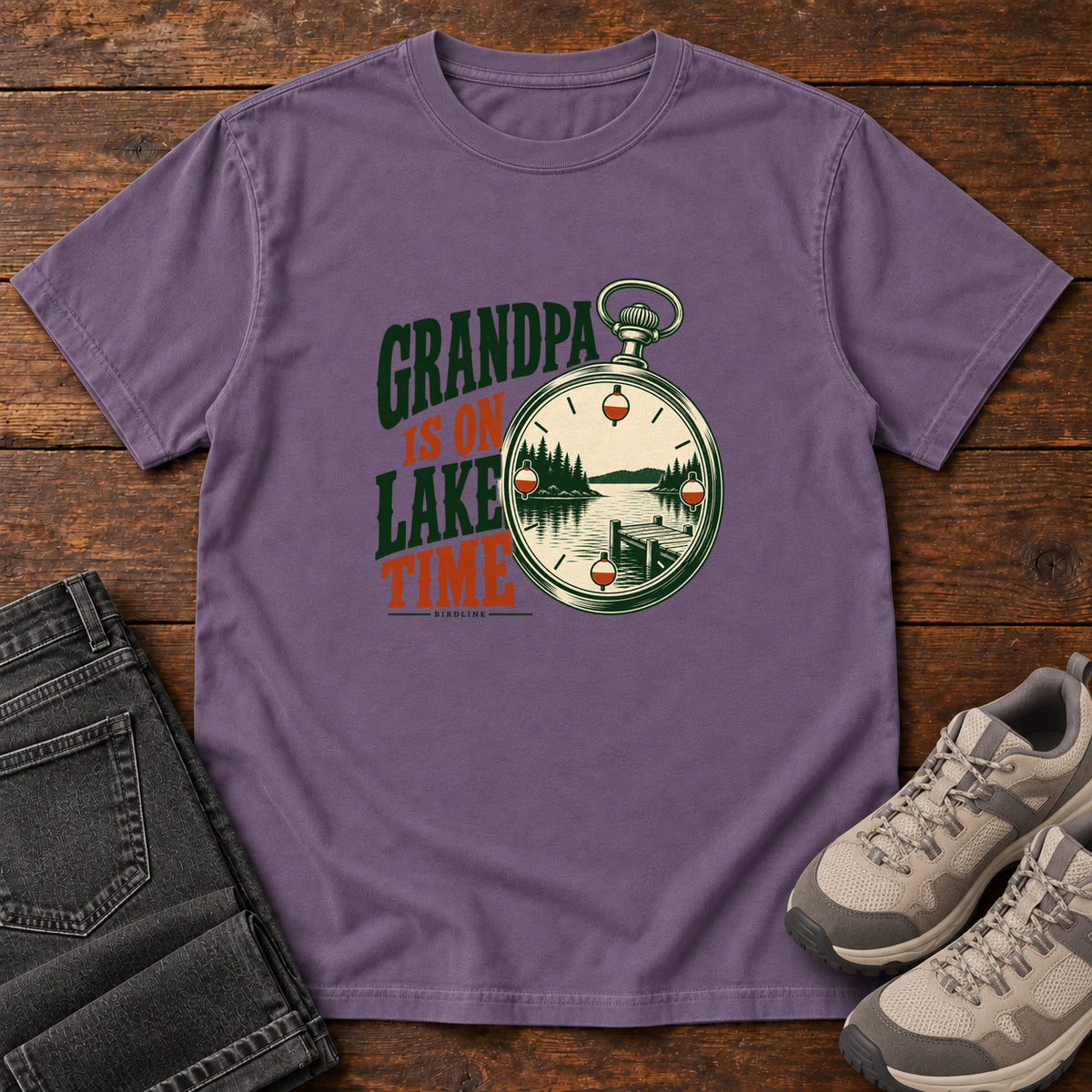 Grape Grandpa Is on Lake Time T-Shirt with a pocket watch framing a lake and dock beside green and orange lettering; front flat lay on wood.