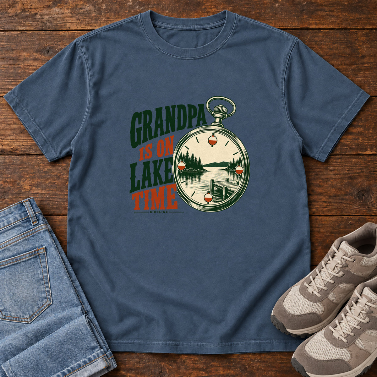 Midnight Grandpa Is on Lake Time T-Shirt with a pocket watch framing a lake and dock beside green and orange lettering; front flat lay on wood.