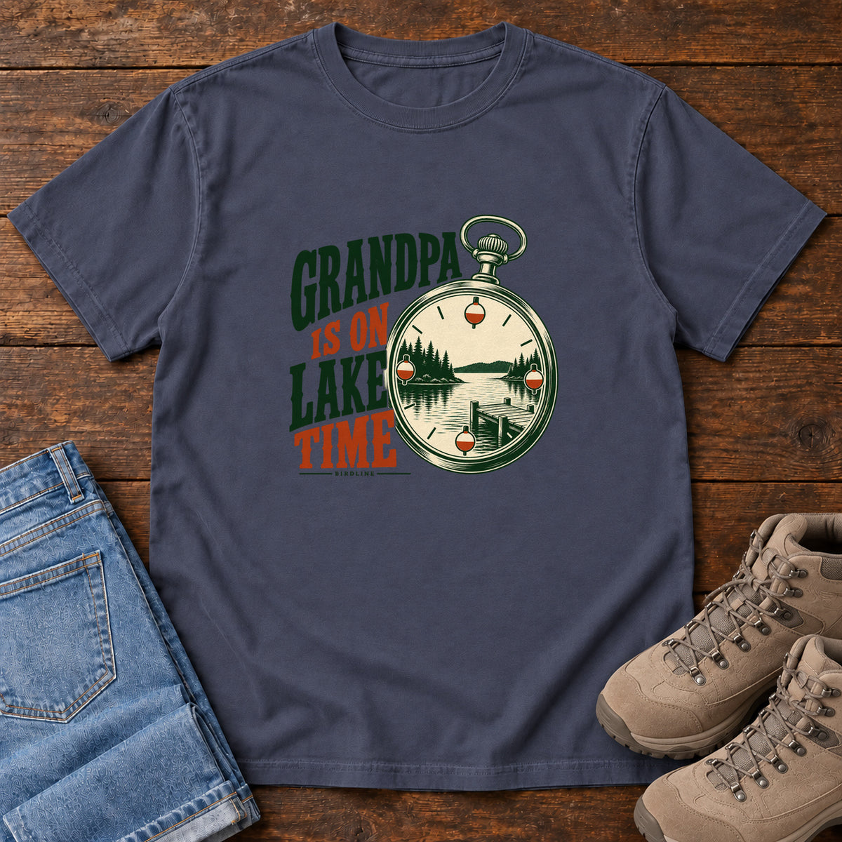 Navy Grandpa Is on Lake Time T-Shirt with a pocket watch framing a lake and dock beside green and orange lettering; front flat lay on wood.