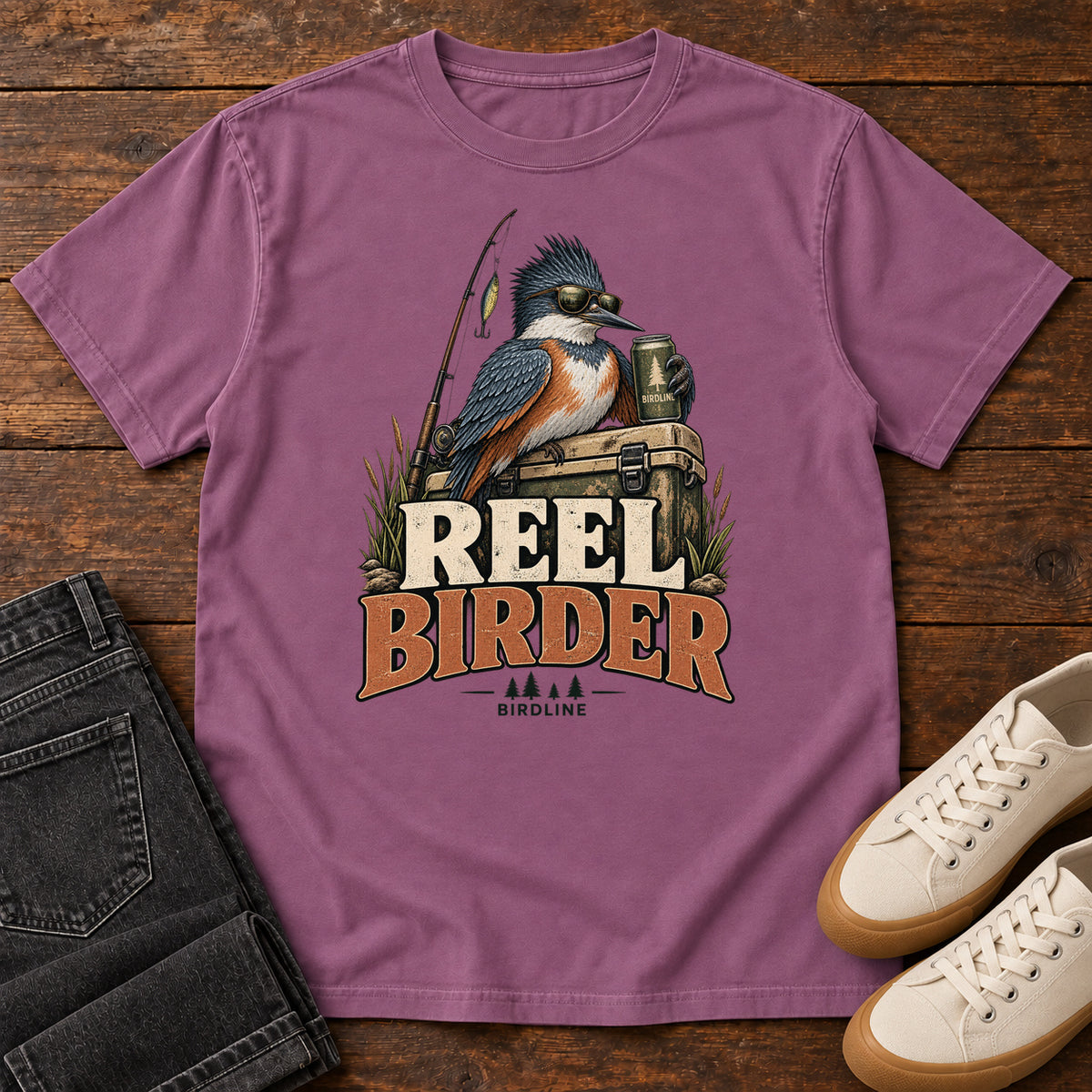Berry Reel Birder T-Shirt with a crested bird in sunglasses holding a mug beside a fishing rod and tackle box; front flat lay on wood.