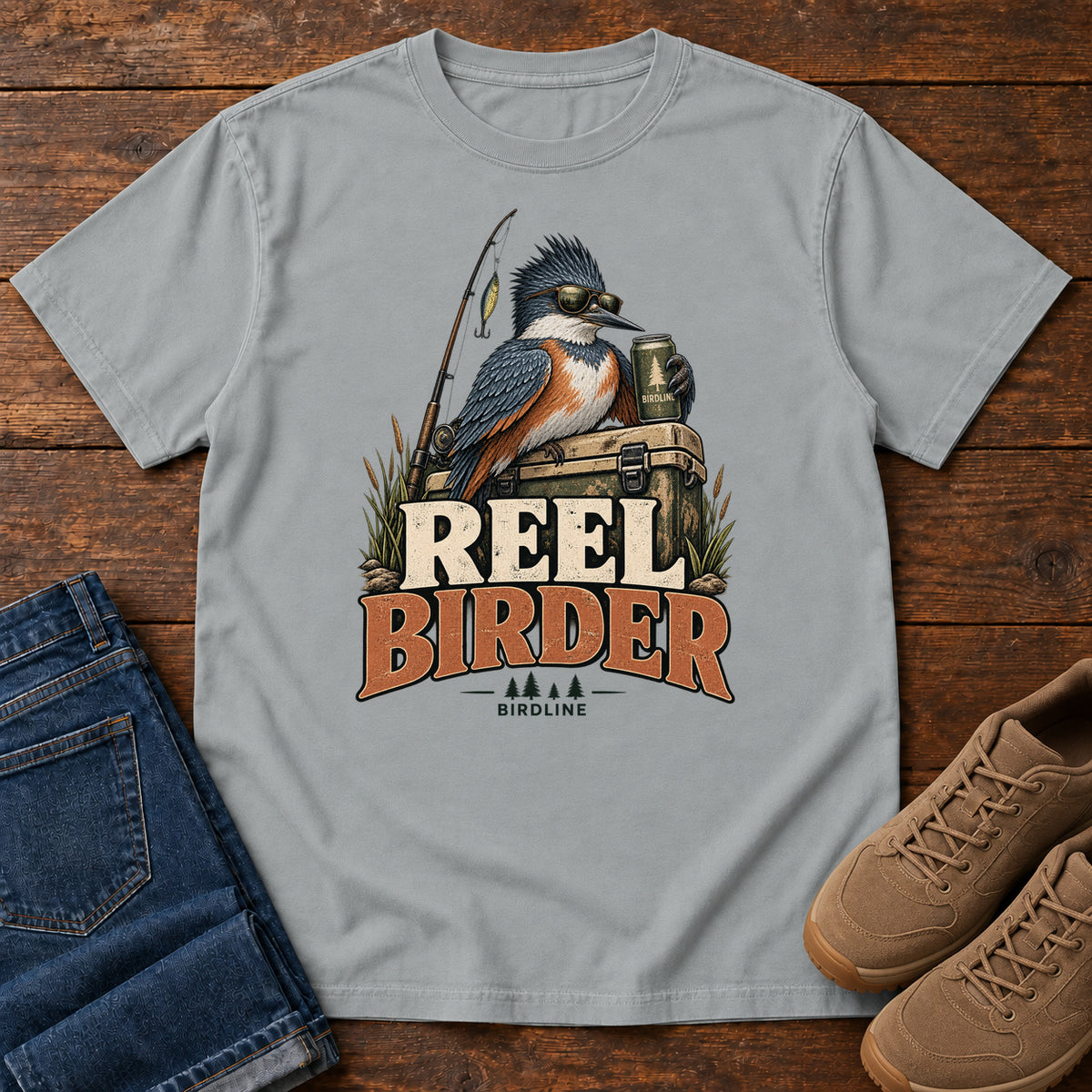 Granite Reel Birder T-Shirt with a crested bird in sunglasses holding a mug beside a fishing rod and tackle box; front flat lay on wood.