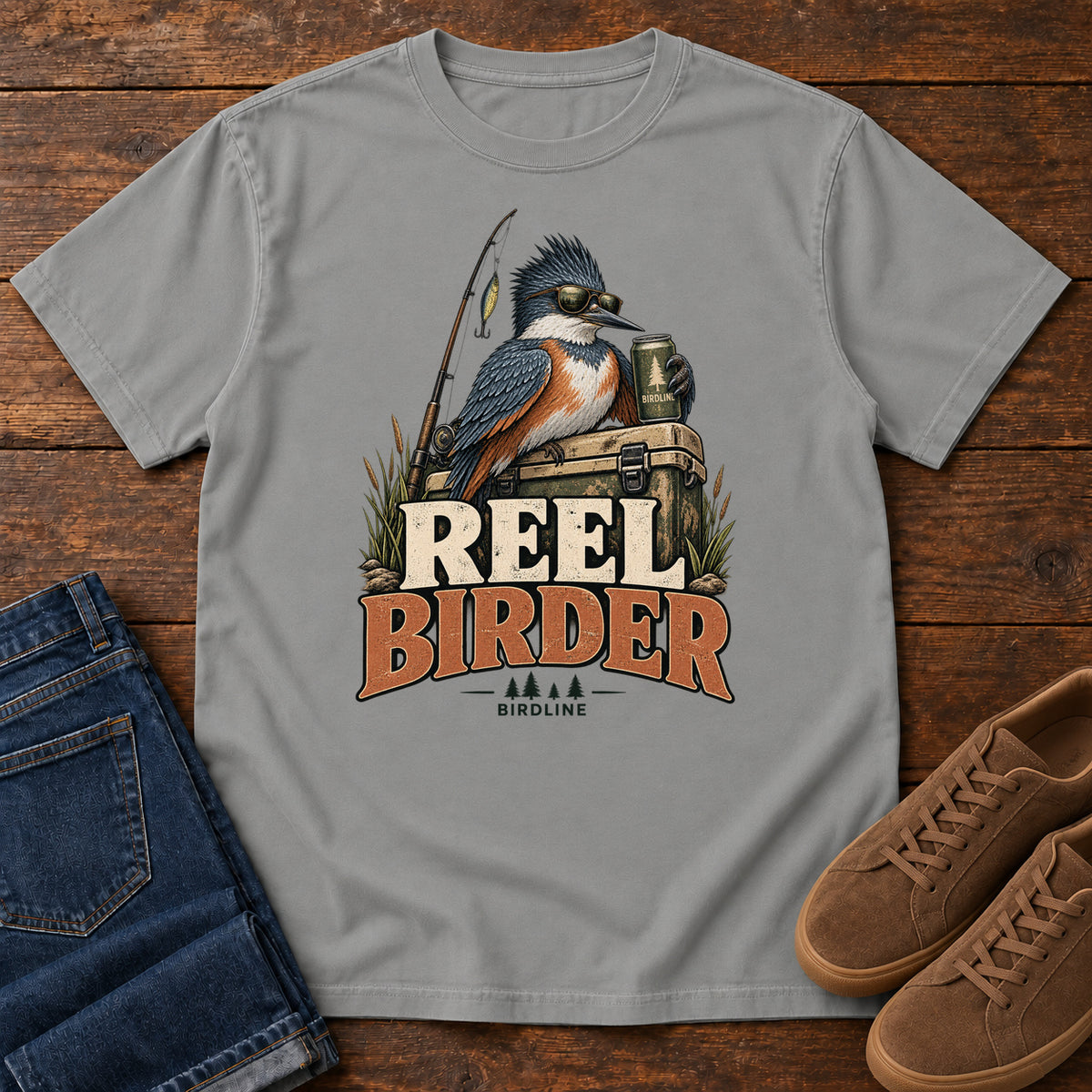 Grey Reel Birder T-Shirt with a crested bird in sunglasses holding a mug beside a fishing rod and tackle box; front flat lay on wood.
