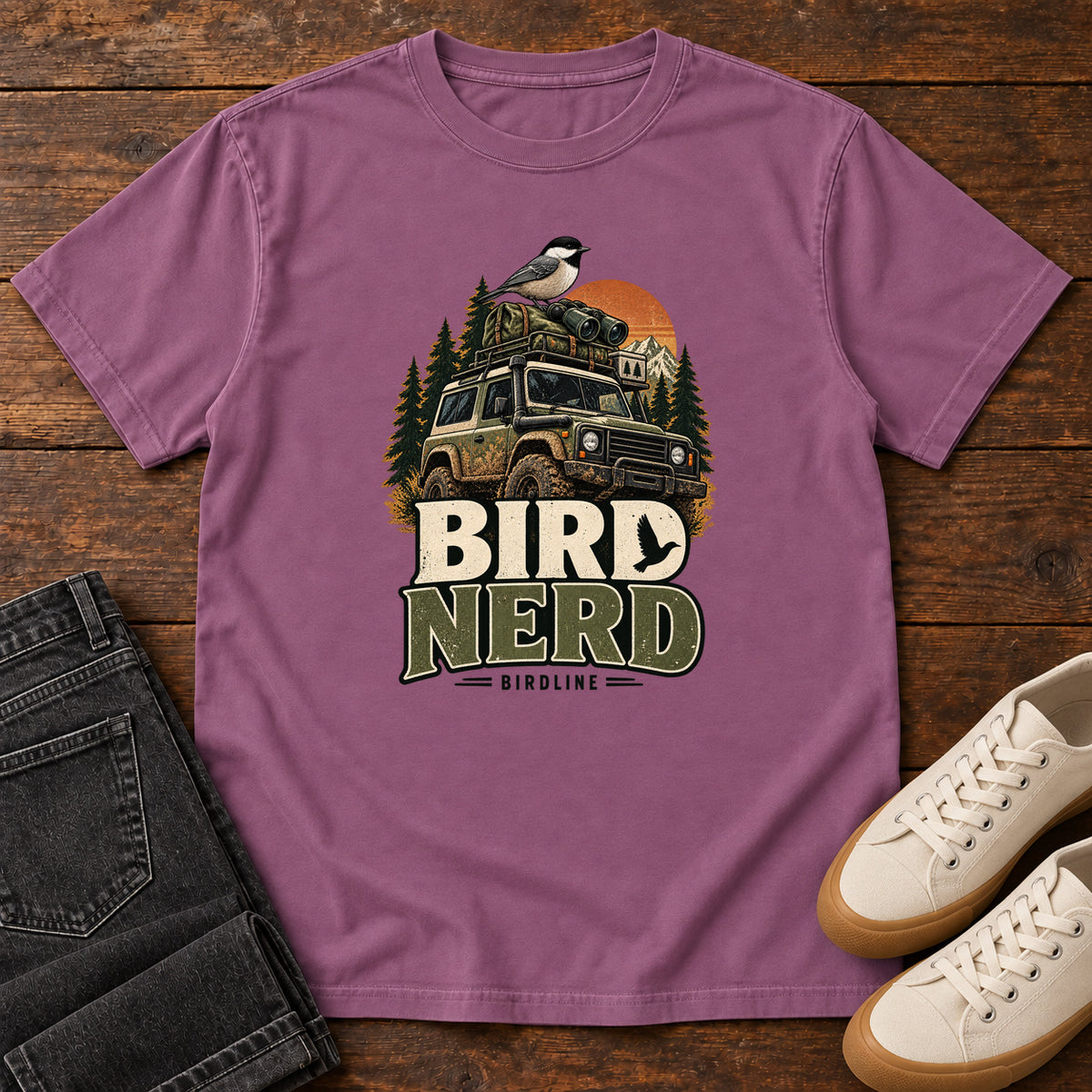 Berry Bird Nerd Off-Roader T-Shirt with a small black-capped bird atop an off-road vehicle, trees and sunset above Bird Nerd lettering, front flat lay on wood.