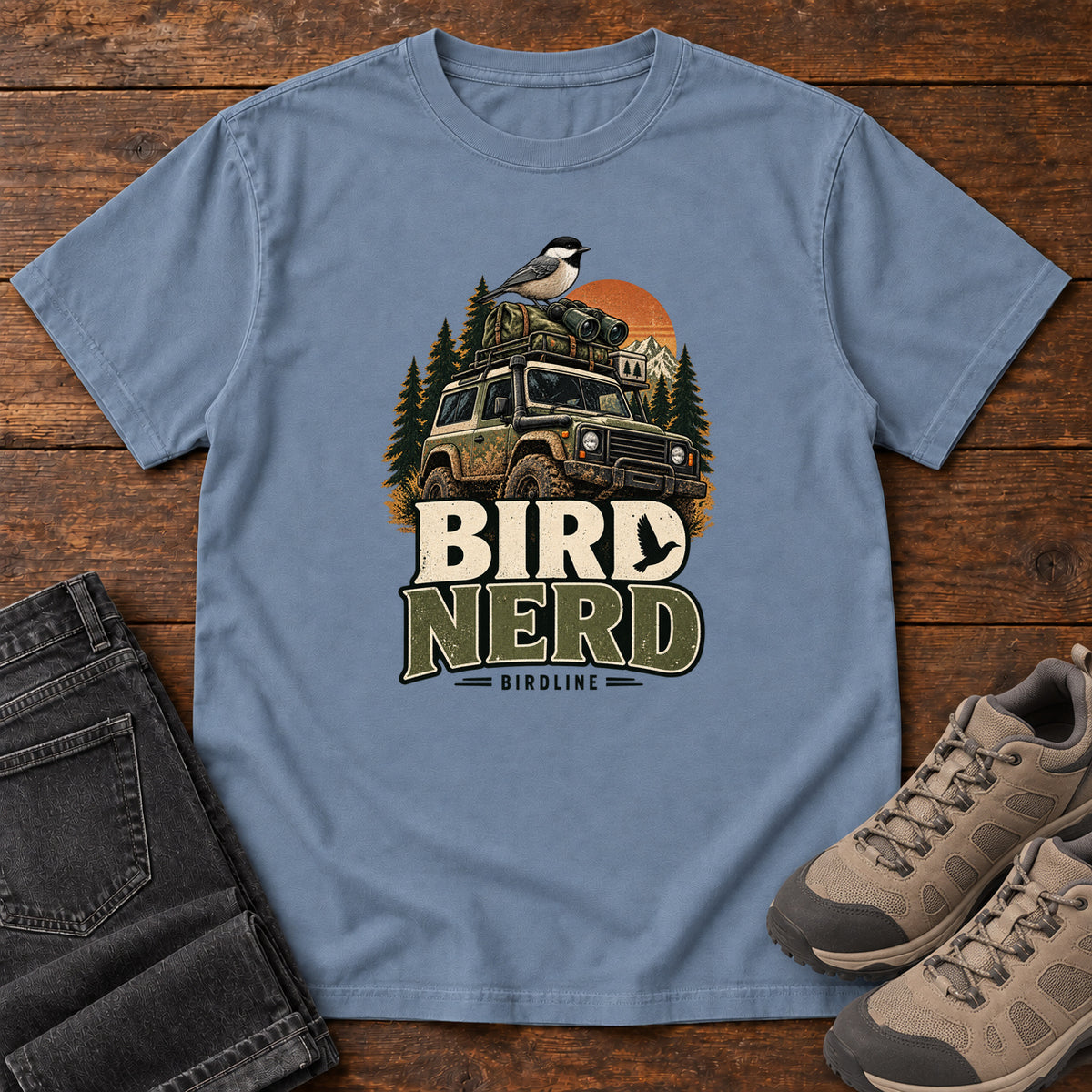 Blue Jean Bird Nerd Off-Roader T-Shirt with a small black-capped bird atop an off-road vehicle, trees and sunset above Bird Nerd lettering, front flat lay on wood.