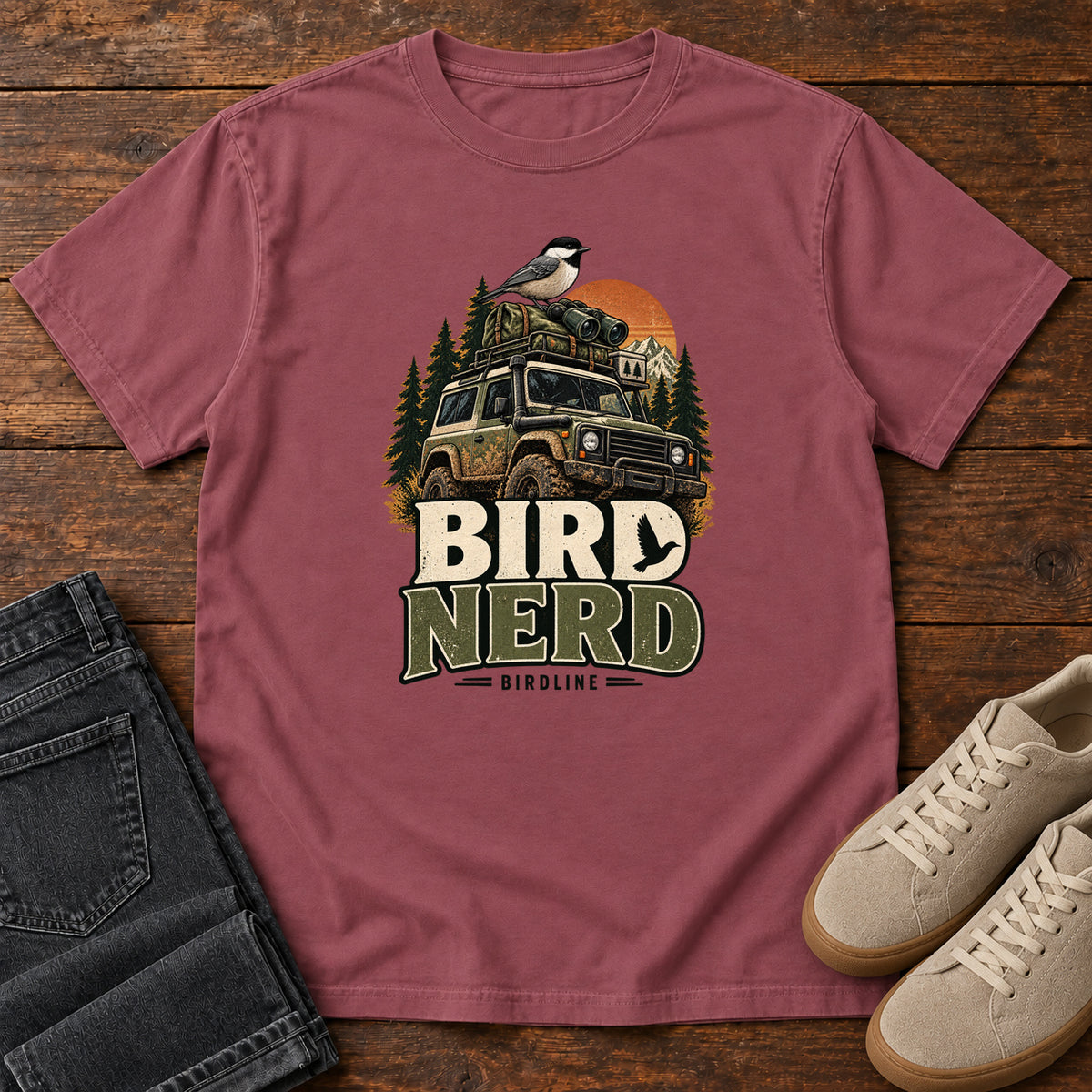 Brick Bird Nerd Off-Roader T-Shirt with a small black-capped bird atop an off-road vehicle, trees and sunset above Bird Nerd lettering, front flat lay on wood.