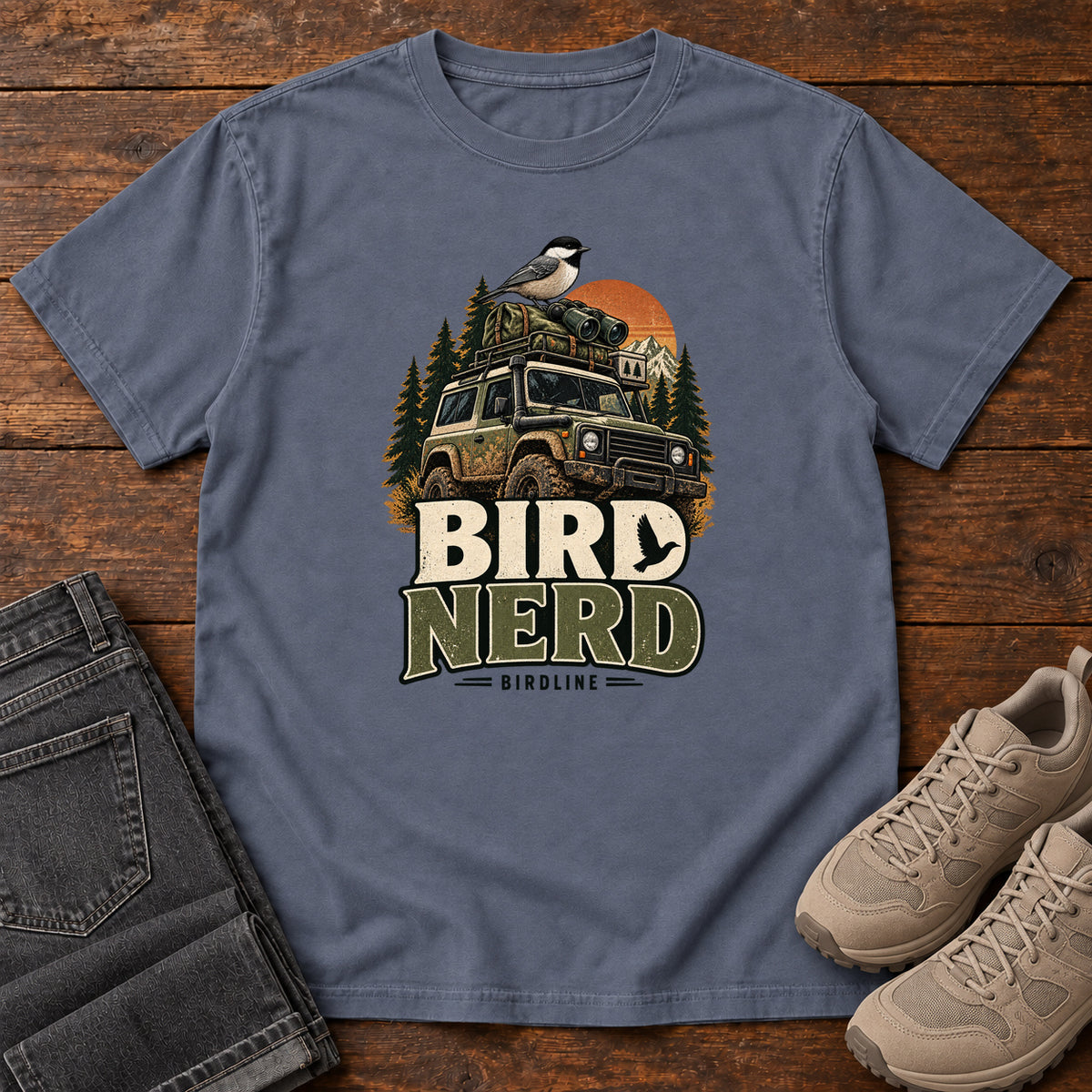 Denim Bird Nerd Off-Roader T-Shirt with a small black-capped bird atop an off-road vehicle, trees and sunset above Bird Nerd lettering, front flat lay on wood.