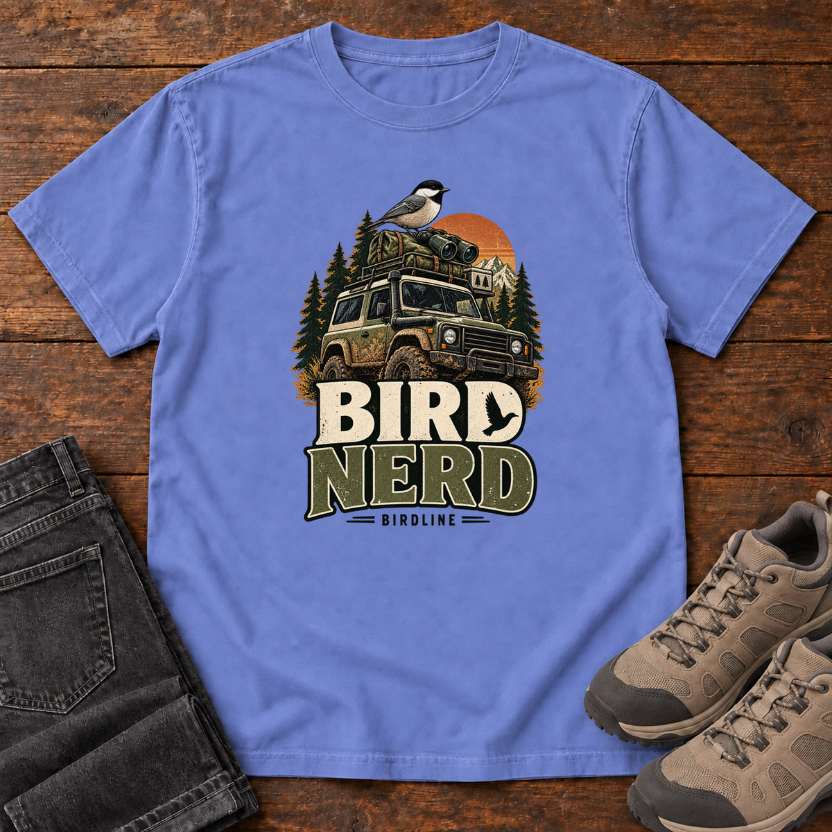 Flo Blue Bird Nerd Off-Roader T-Shirt with a small black-capped bird atop an off-road vehicle, trees and sunset above Bird Nerd lettering, front flat lay on wood.