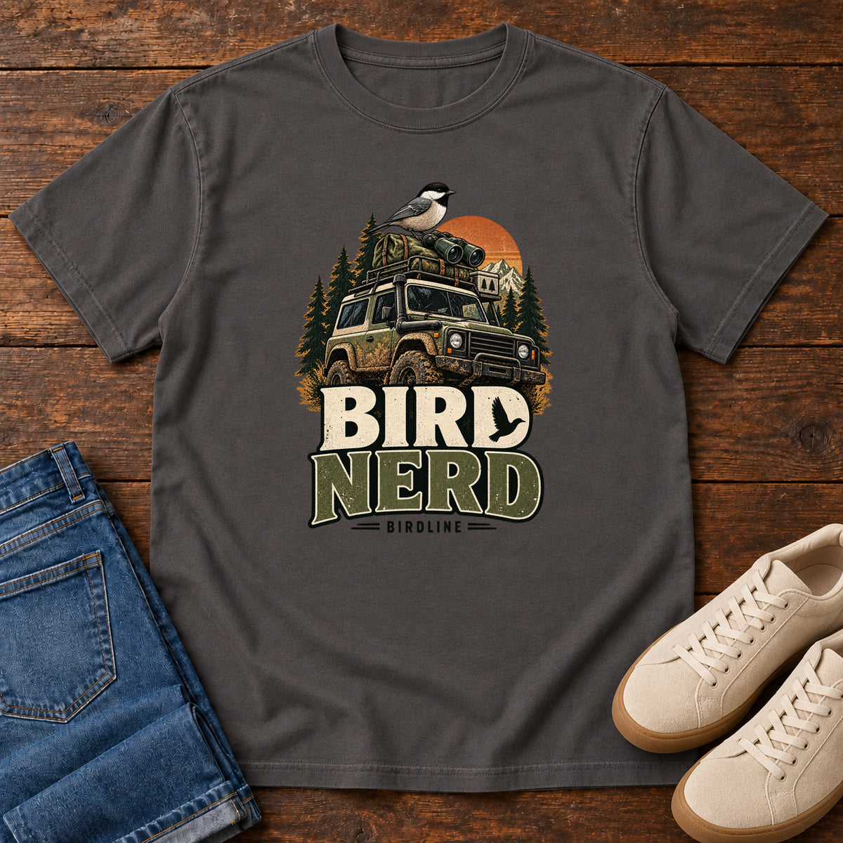 Graphite Bird Nerd Off-Roader T-Shirt with a small black-capped bird atop an off-road vehicle, trees and sunset above Bird Nerd lettering, front flat lay on wood.