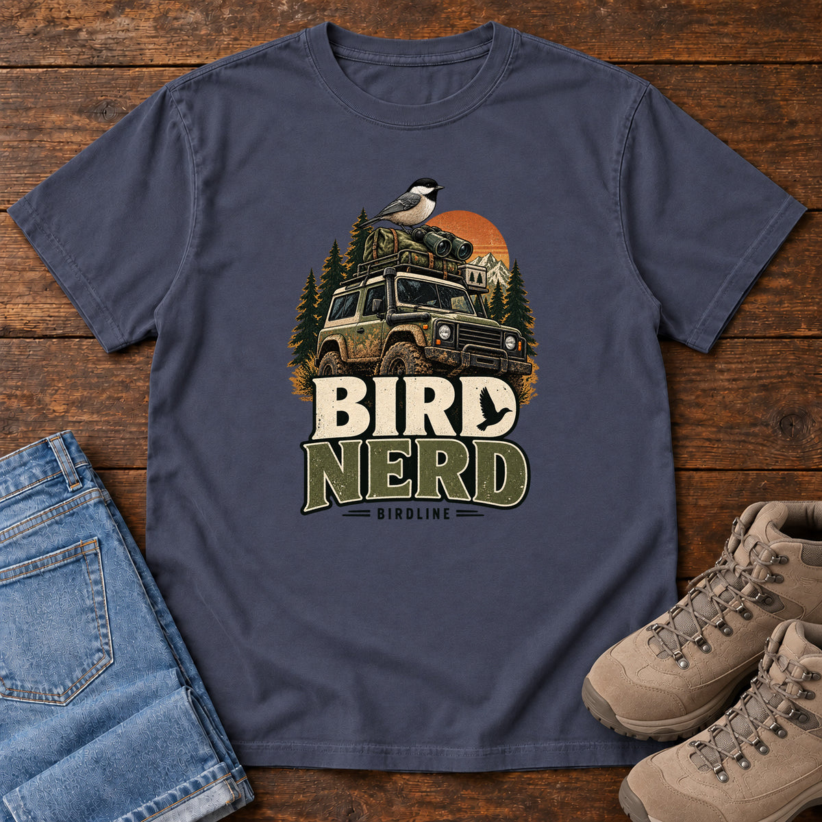 Navy Bird Nerd Off-Roader T-Shirt with a small black-capped bird atop an off-road vehicle, trees and sunset above Bird Nerd lettering, front flat lay on wood.