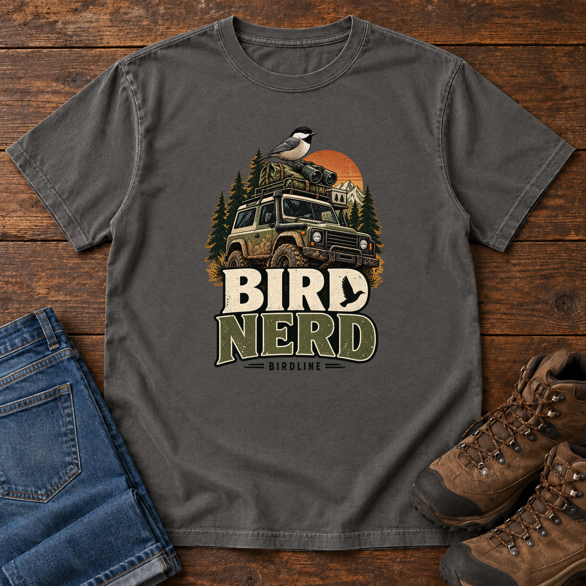Pepper Bird Nerd Off-Roader T-Shirt with a small black-capped bird atop an off-road vehicle, trees and sunset above Bird Nerd lettering, front flat lay on wood.