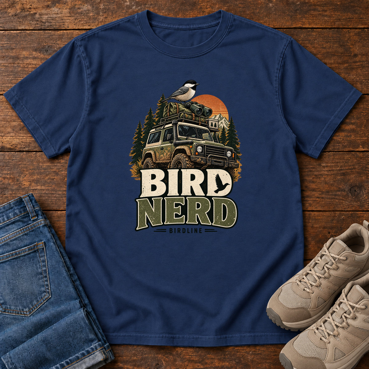 True Navy Bird Nerd Off-Roader T-Shirt with a small black-capped bird atop an off-road vehicle, trees and sunset above Bird Nerd lettering, front flat lay on wood.