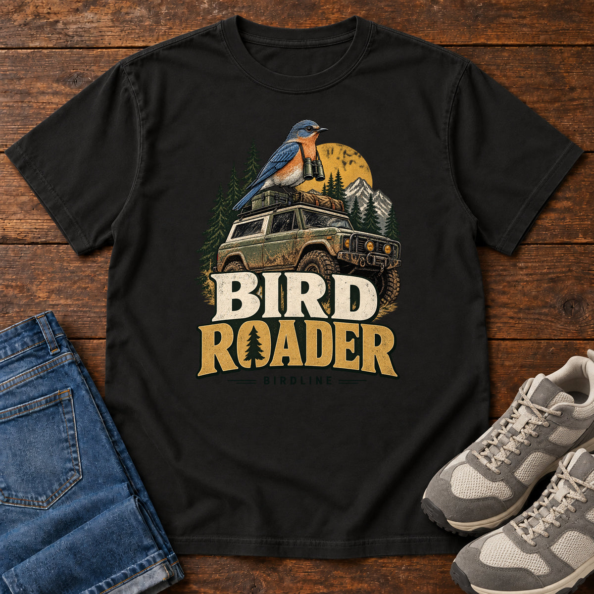 Bird Roader T-Shirt in Black, front laid flat on wood, featuring a blue-and-orange bird above an off-road vehicle, pine trees, mountains and a yellow sun.