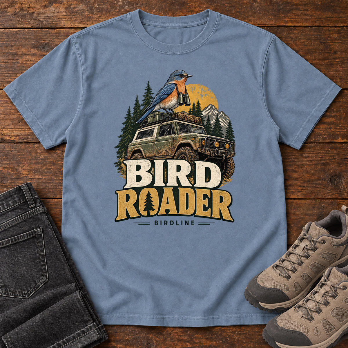Bird Roader T-Shirt in Blue Jean, front laid flat on wood, featuring a blue-and-orange bird above an off-road vehicle, pine trees, mountains and a yellow sun.