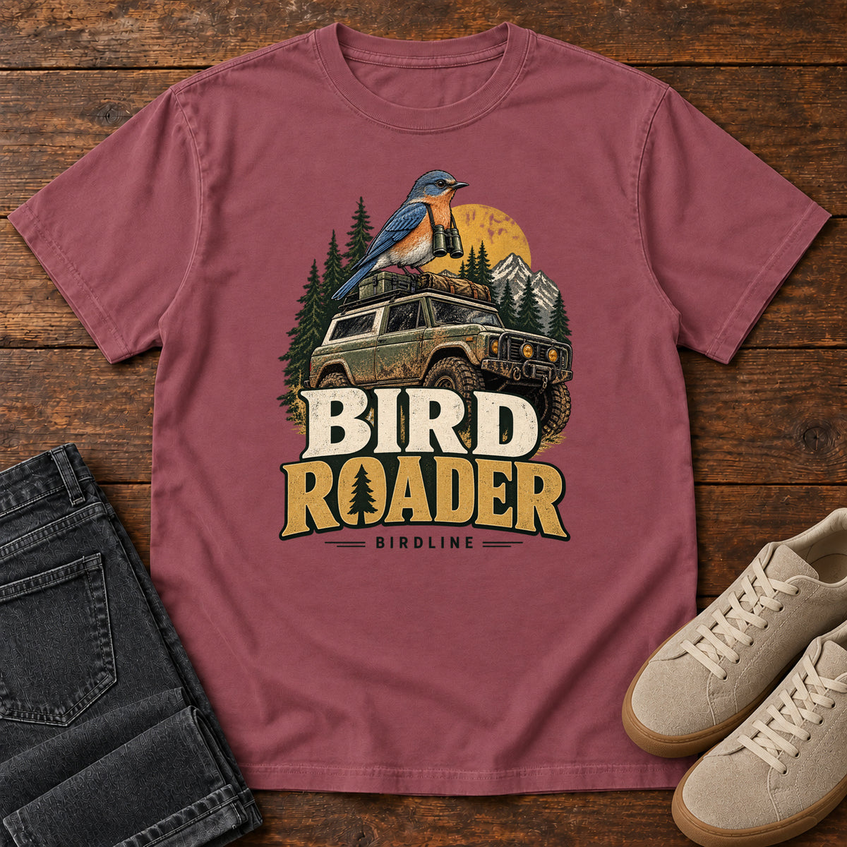 Bird Roader T-Shirt in Brick, front laid flat on wood, featuring a blue-and-orange bird above an off-road vehicle, pine trees, mountains and a yellow sun.