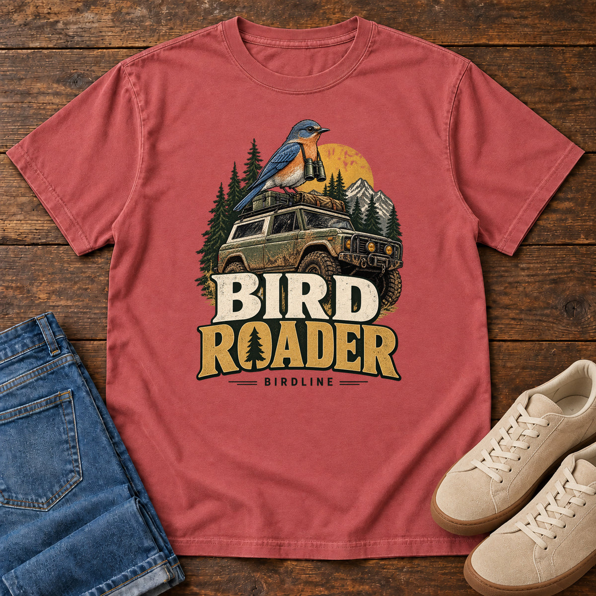 Bird Roader T-Shirt in Crimson, front laid flat on wood, featuring a blue-and-orange bird above an off-road vehicle, pine trees, mountains and a yellow sun.