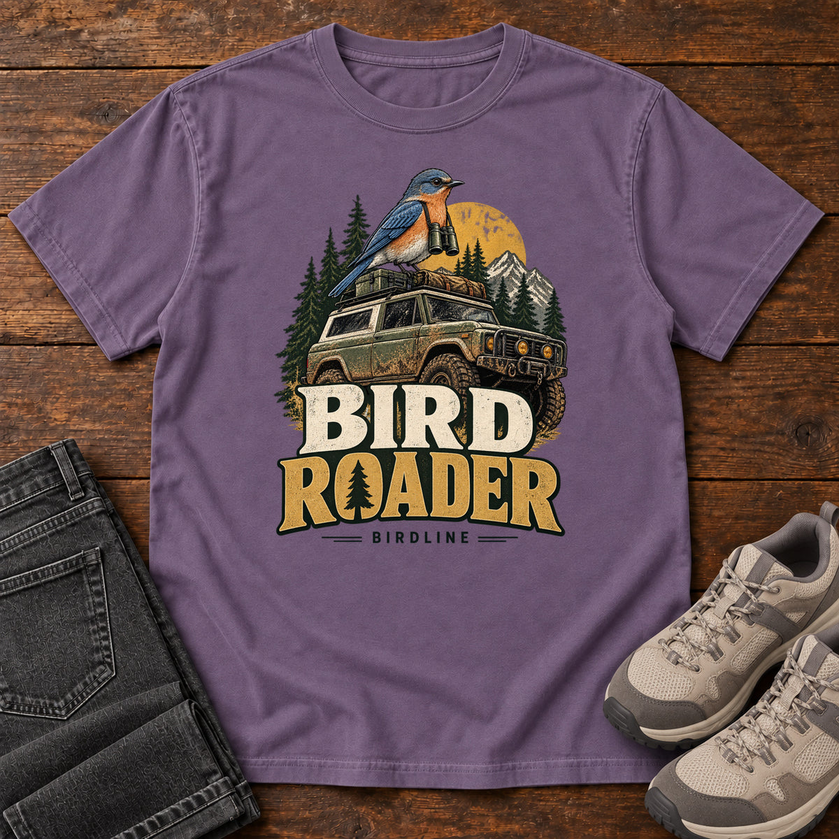 Bird Roader T-Shirt in Grape, front laid flat on wood, featuring a blue-and-orange bird above an off-road vehicle, pine trees, mountains and a yellow sun.