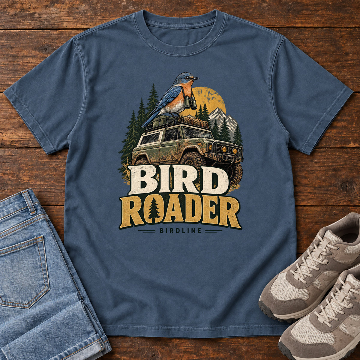 Bird Roader T-Shirt in Midnight, front laid flat on wood, featuring a blue-and-orange bird above an off-road vehicle, pine trees, mountains and a yellow sun.