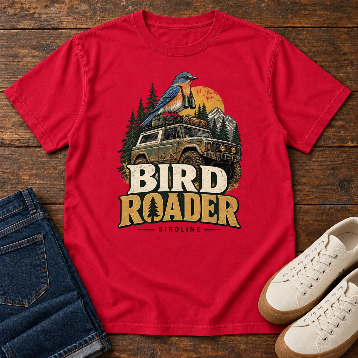 Bird Roader T-Shirt in Red, front laid flat on wood, featuring a blue-and-orange bird above an off-road vehicle, pine trees, mountains and a yellow sun.