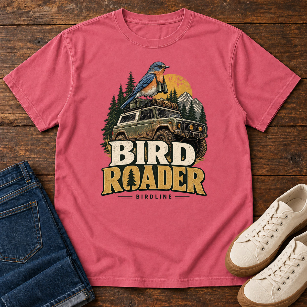 Bird Roader T-Shirt in Watermelon, front laid flat on wood, featuring a blue-and-orange bird above an off-road vehicle, pine trees, mountains and a yellow sun.