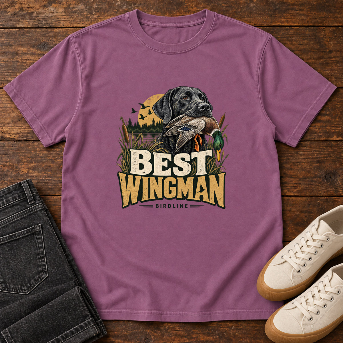 Berry Best Wingman T-Shirt with a black dog carrying a duck, marsh reeds and cream-and-gold lettering, front flat lay on wood.