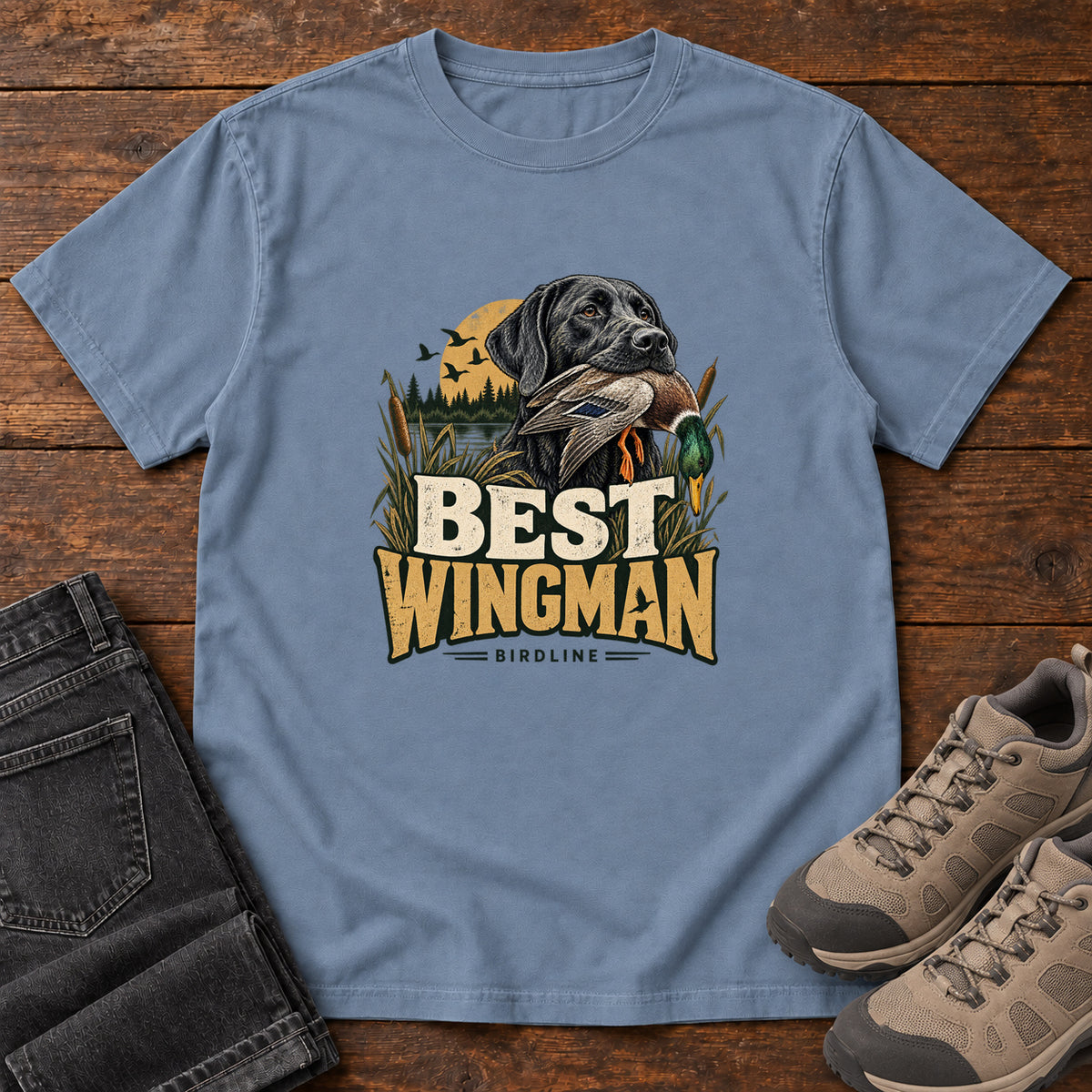 Blue Jean Best Wingman T-Shirt with a black dog carrying a duck, marsh reeds and cream-and-gold lettering, front flat lay on wood.