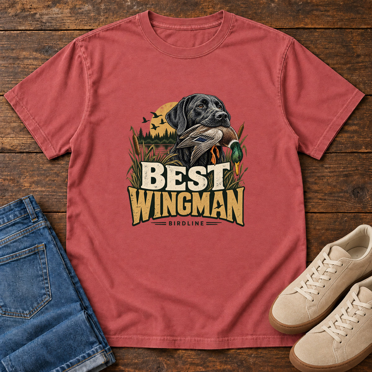 Crimson Best Wingman T-Shirt with a black dog carrying a duck, marsh reeds and cream-and-gold lettering, front flat lay on wood.