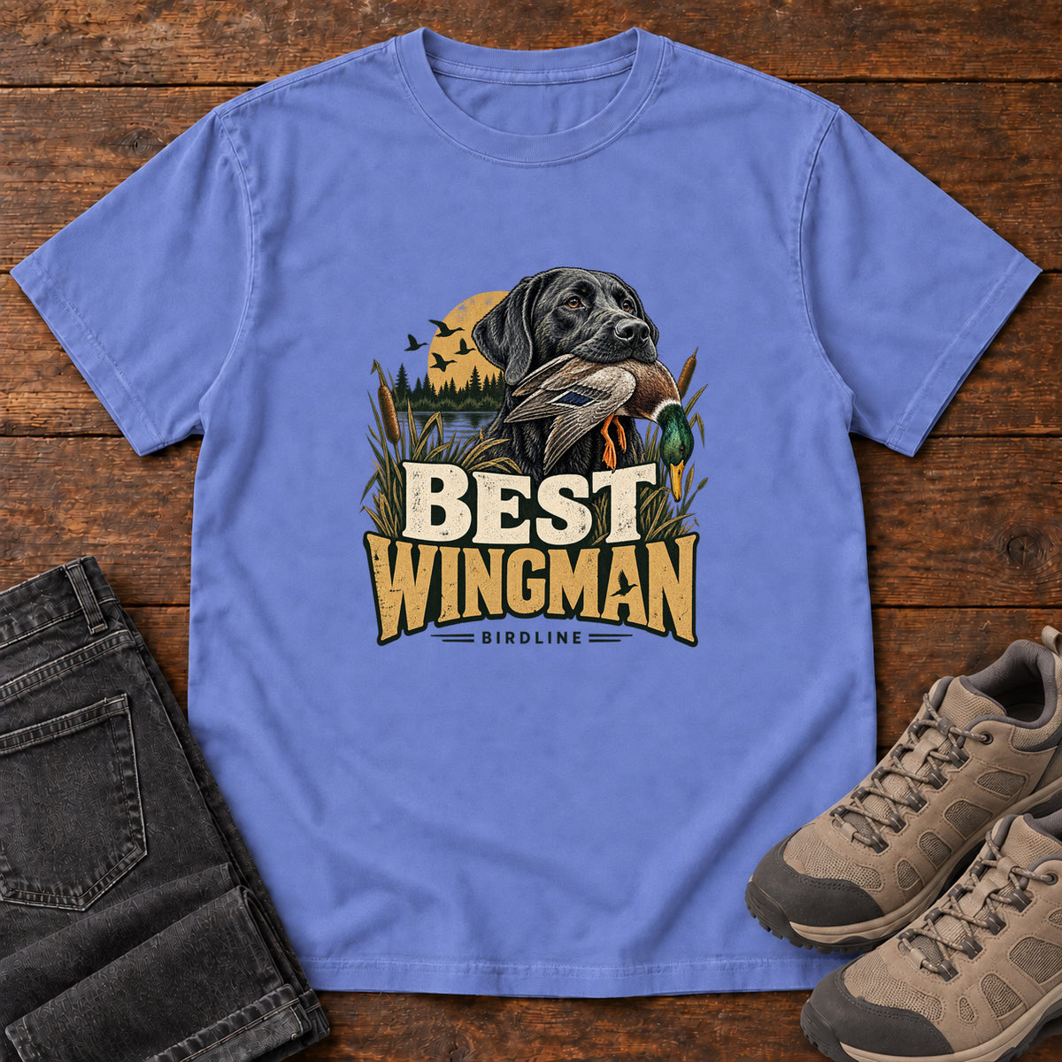 Flo Blue Best Wingman T-Shirt with a black dog carrying a duck, marsh reeds and cream-and-gold lettering, front flat lay on wood.