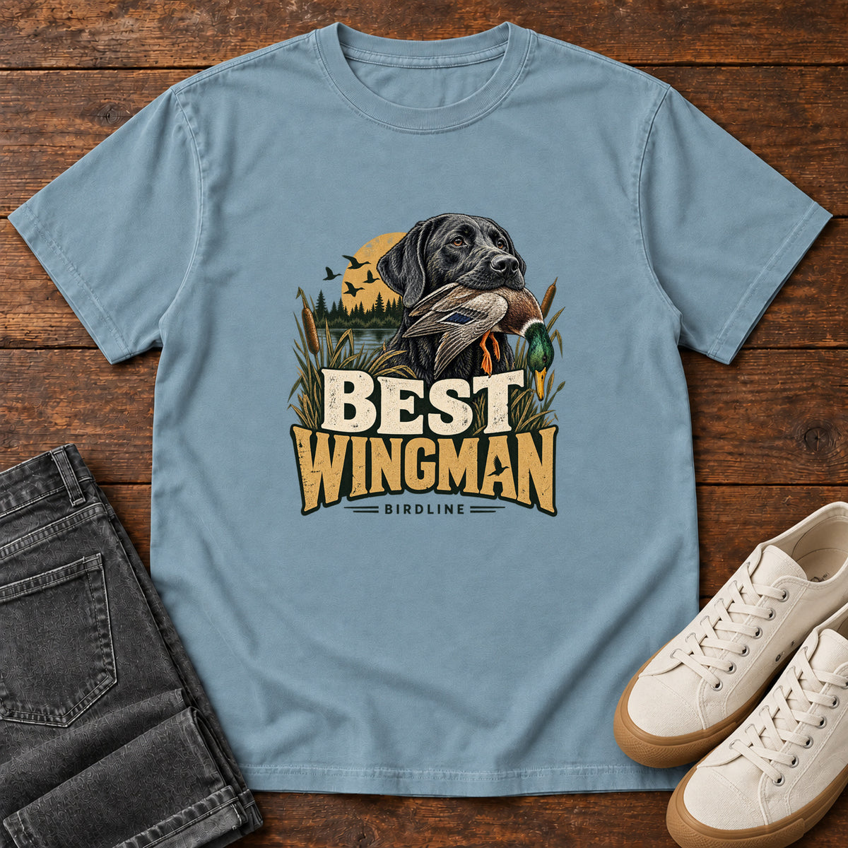 Ice Blue Best Wingman T-Shirt with a black dog carrying a duck, marsh reeds and cream-and-gold lettering, front flat lay on wood.