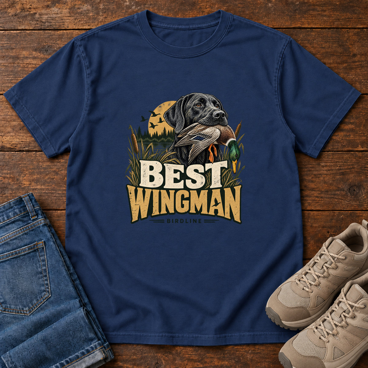 True Navy Best Wingman T-Shirt with a black dog carrying a duck, marsh reeds and cream-and-gold lettering, front flat lay on wood.
