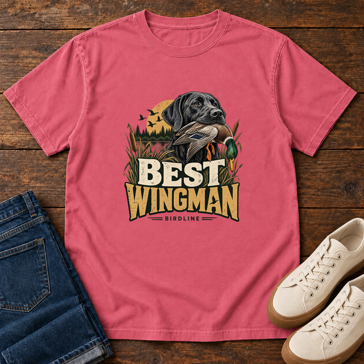 Watermelon Best Wingman T-Shirt with a black dog carrying a duck, marsh reeds and cream-and-gold lettering, front flat lay on wood.
