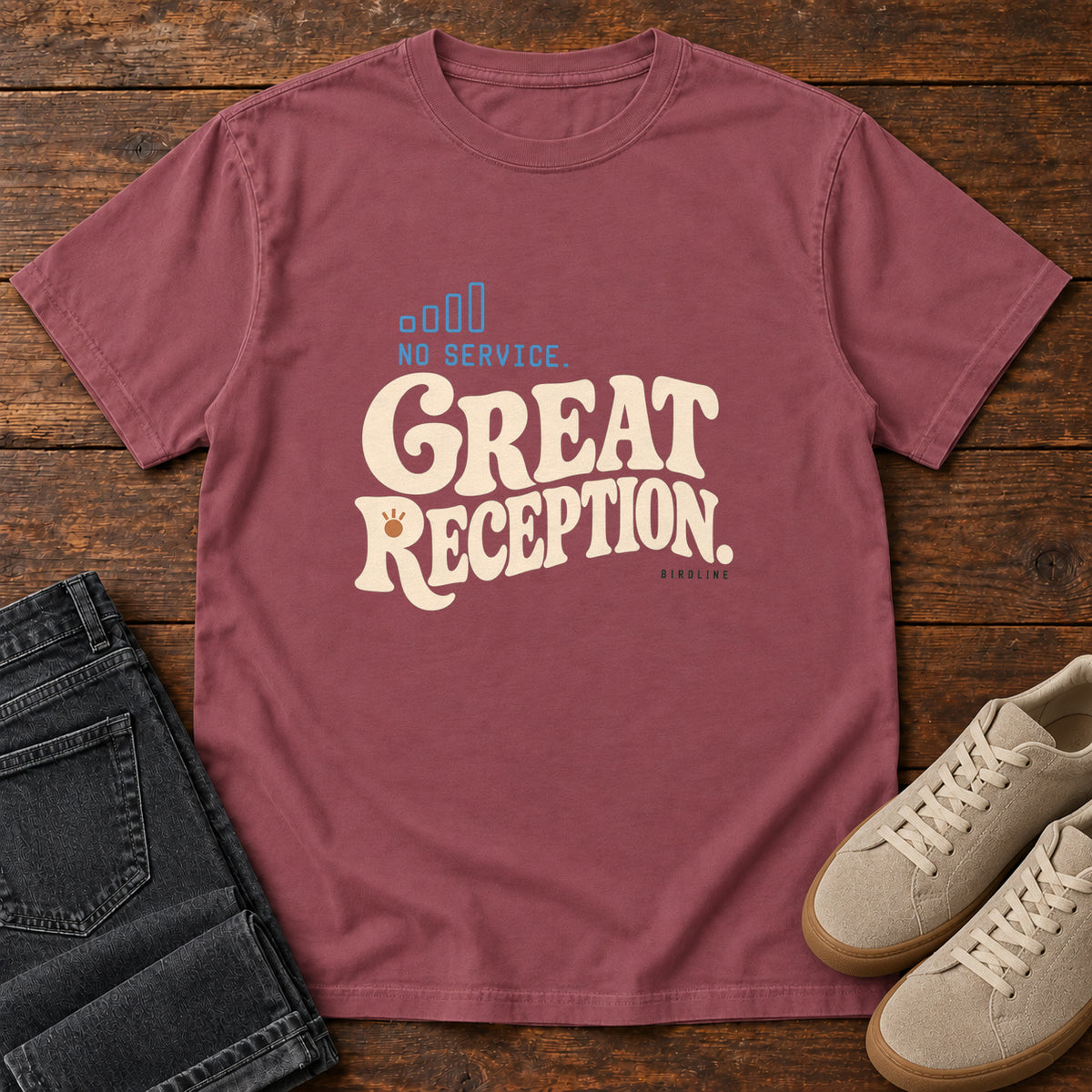 Brick No Service. Great Reception. T-Shirt with cream retro lettering and blue outlined phone-signal bars; front flat lay on wood.