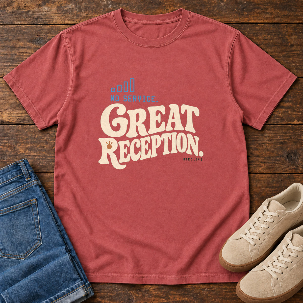 Crimson No Service. Great Reception. T-Shirt with cream retro lettering and blue outlined phone-signal bars; front flat lay on wood.