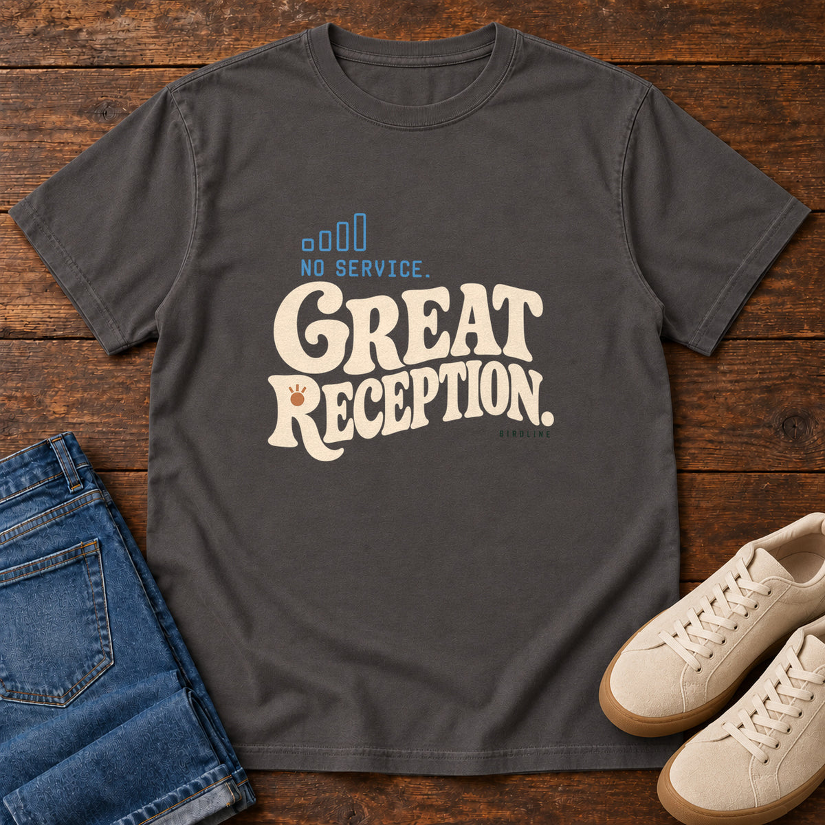 Graphite No Service. Great Reception. T-Shirt with cream retro lettering and blue outlined phone-signal bars; front flat lay on wood.