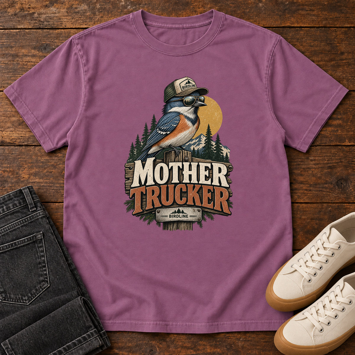 Berry Mother Trucker T-Shirt with a blue-and-orange bird wearing a cap and sunglasses above mountains and pines; front flat lay on wood.