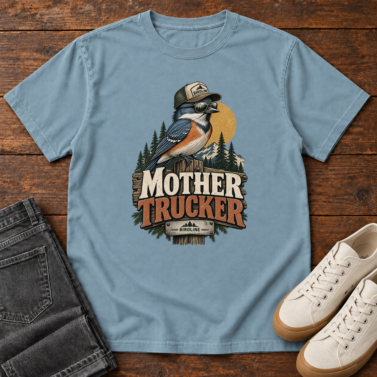 Ice Blue Mother Trucker T-Shirt with a blue-and-orange bird wearing a cap and sunglasses above mountains and pines; front flat lay on wood.