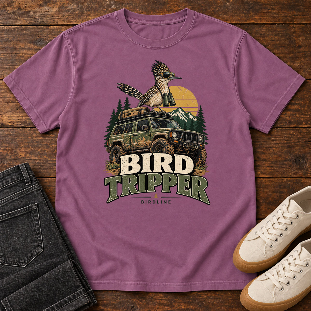 Berry Bird Tripper T-Shirt with a crested, long-tailed bird with binoculars above an off-road vehicle and Bird Tripper lettering, front flat lay on wood.
