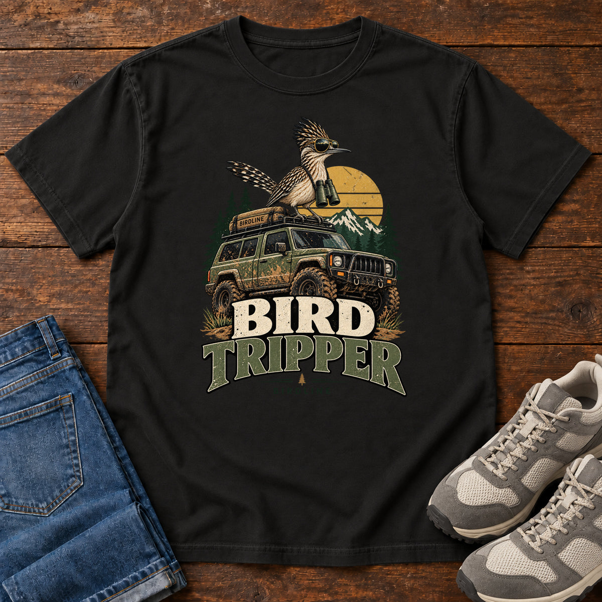 Black Bird Tripper T-Shirt with a crested, long-tailed bird with binoculars above an off-road vehicle and Bird Tripper lettering, front flat lay on wood.