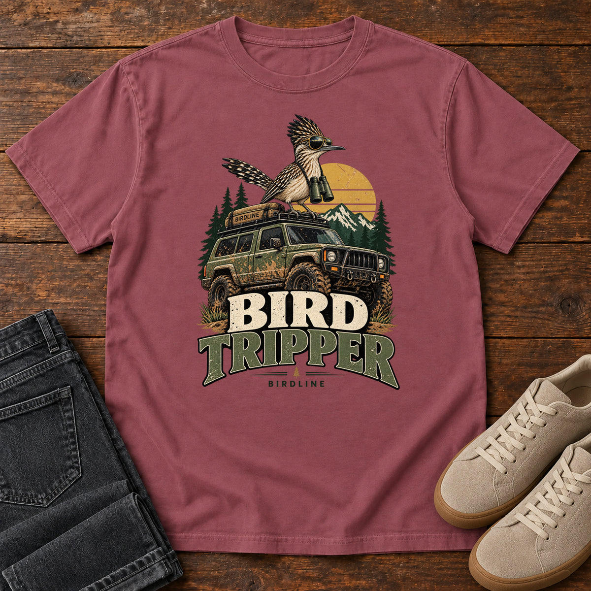 Brick Bird Tripper T-Shirt with a crested, long-tailed bird with binoculars above an off-road vehicle and Bird Tripper lettering, front flat lay on wood.