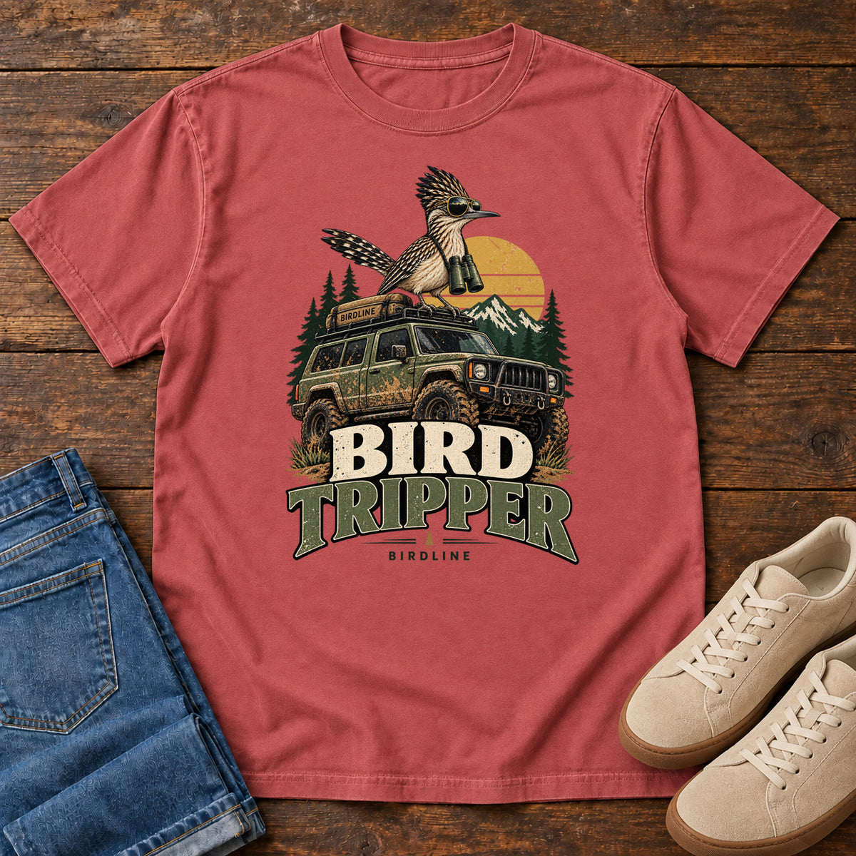 Crimson Bird Tripper T-Shirt with a crested, long-tailed bird with binoculars above an off-road vehicle and Bird Tripper lettering, front flat lay on wood.