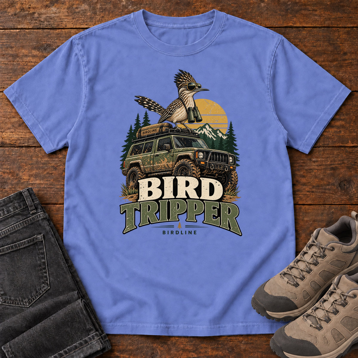 Flo Blue Bird Tripper T-Shirt with a crested, long-tailed bird with binoculars above an off-road vehicle and Bird Tripper lettering, front flat lay on wood.
