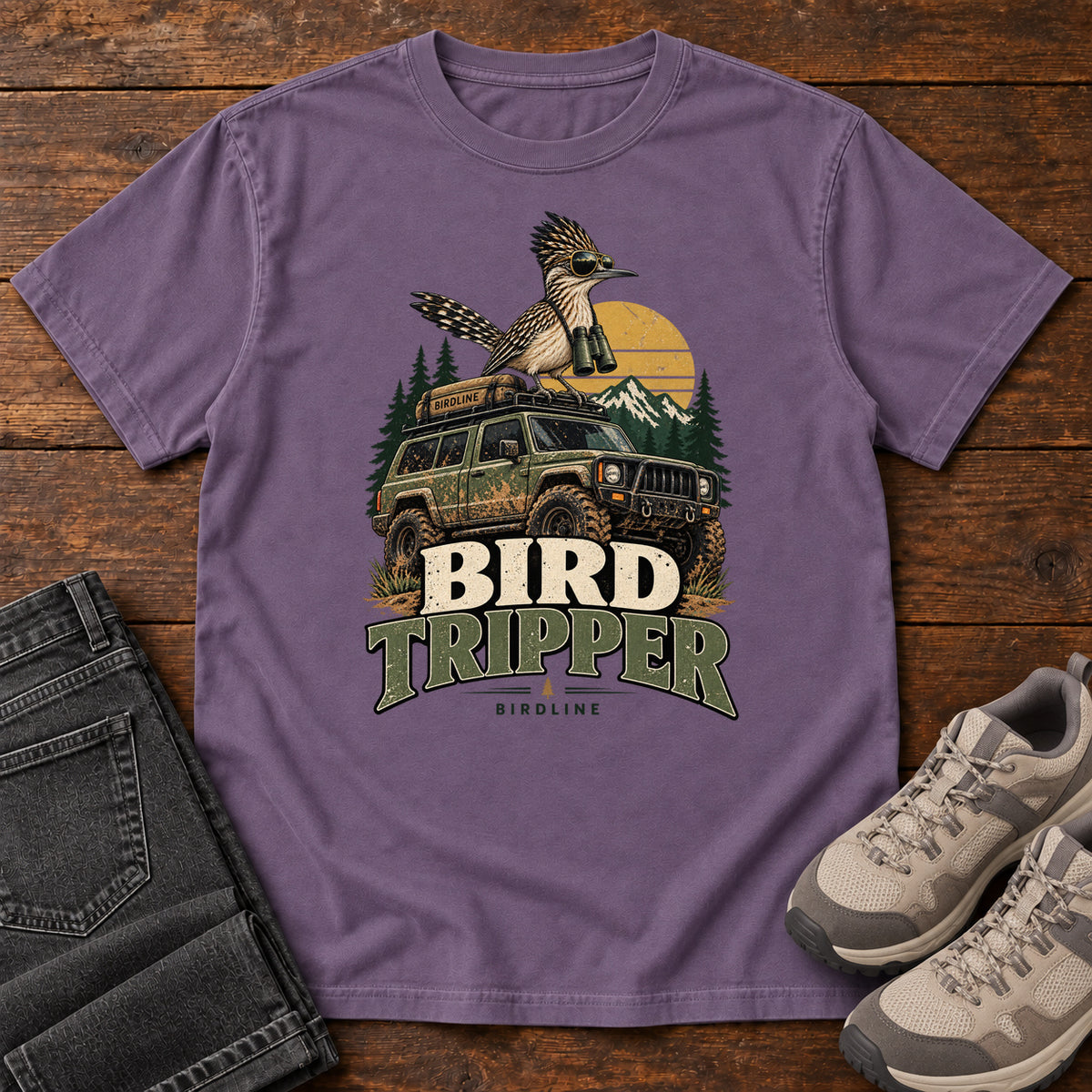 Grape Bird Tripper T-Shirt with a crested, long-tailed bird with binoculars above an off-road vehicle and Bird Tripper lettering, front flat lay on wood.